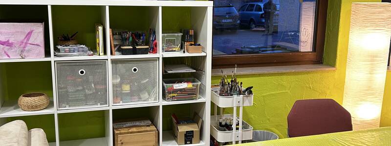 Ein heller Raum mit gelb gestrichenen Wänden zeigt ein Kreativstudio mit Regalen voller Kunstmaterialien, mehreren Stühlen und einem großen Tisch.