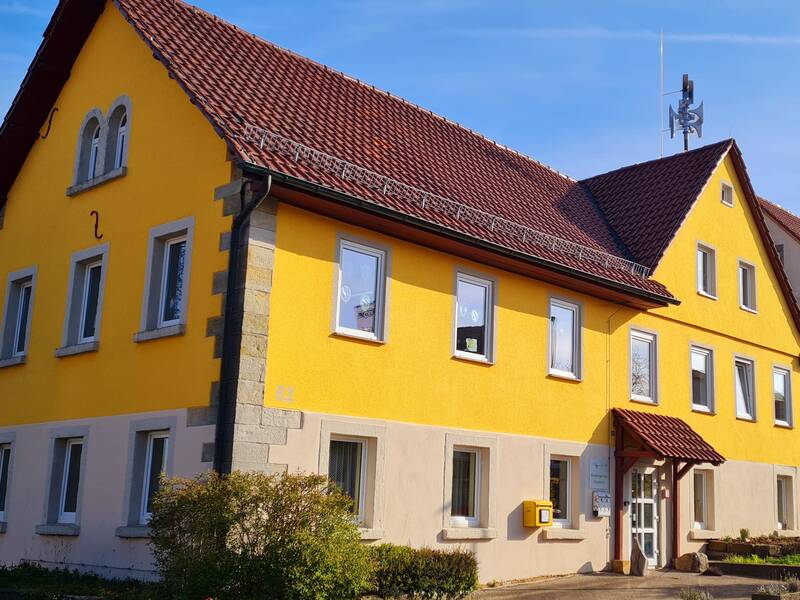 Das Bild zeigt ein zweistöckiges gelb- und beigefarbenes Haus mit einem roten Ziegeldach und mehreren Fenstern, vor dem ein kleiner Busch wächst.