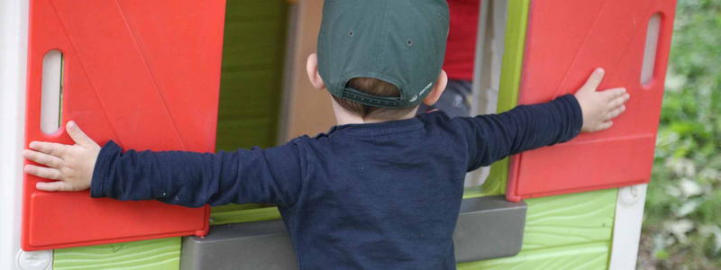 Ein Kind von hinten welches die Arme ganz ausgebreitet hat und vor einem Spielhaus steht und die Fenster schließen möchte