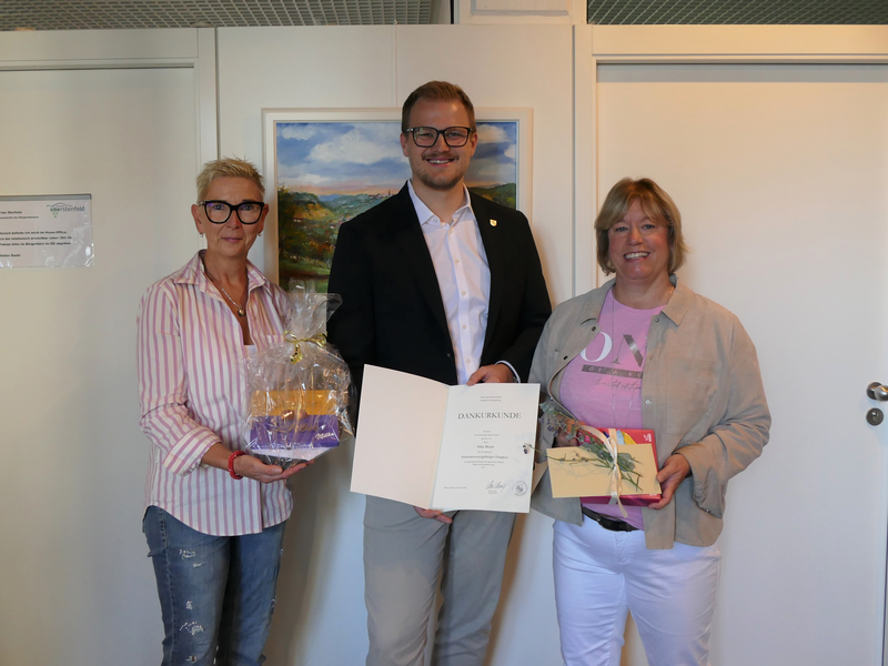 Drei Personen stehen lächelnd nebeneinander, wobei die mittlere Person eine Urkunde hält und die beiden anderen jeweils ein Geschenk präsentieren.