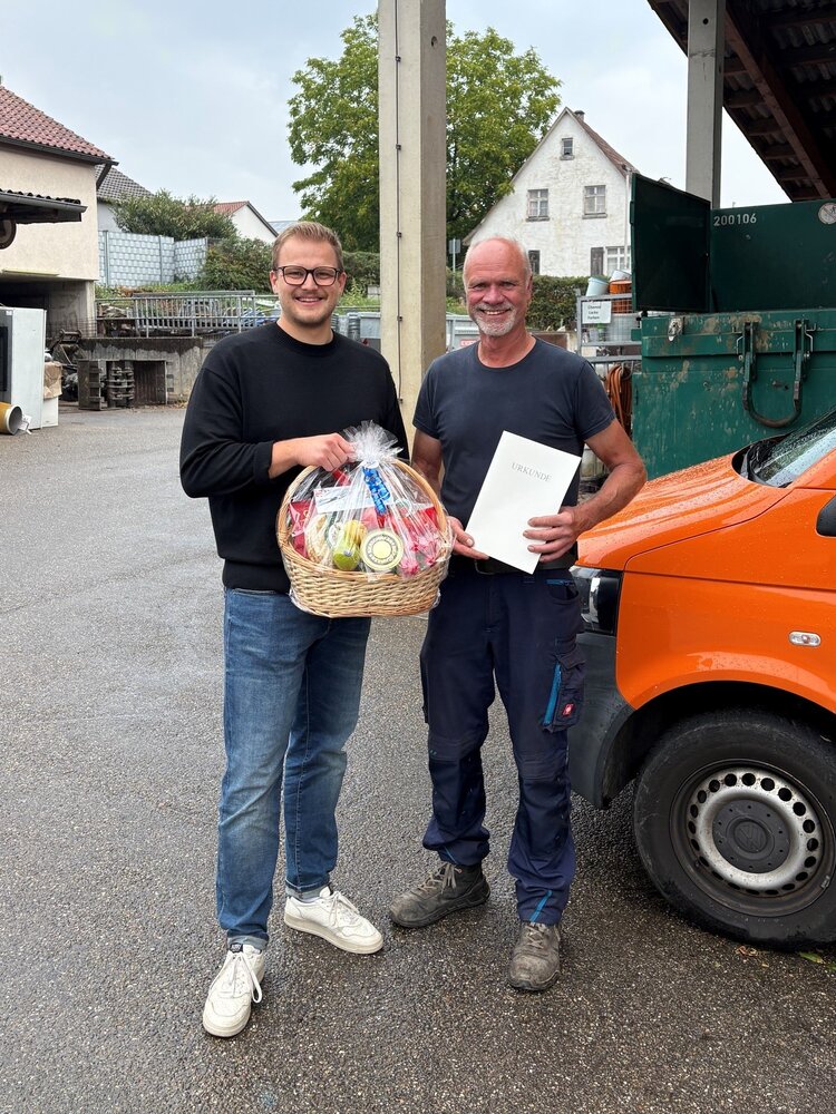 Zwei Männer stehen lächelnd nebeneinander auf einem Hofplatz, einer hält einen Geschenkkorb und der andere eine Urkunde.