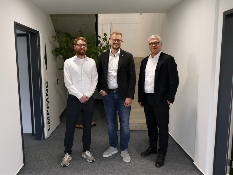 Drei Männer in formeller Kleidung stehen in einem Büroflur vor einer Wand mit dem Wort "Empfang".