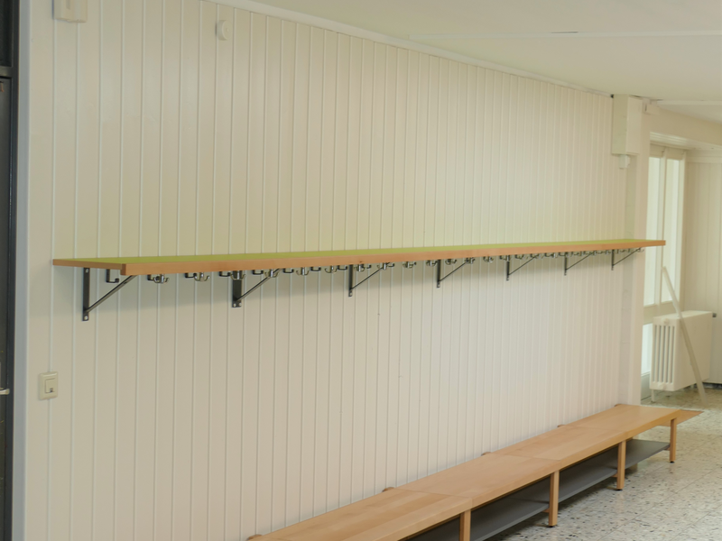 Ein langer Holzbank mit einer darüberliegenden Hutablage und darunter einer Reihe von Haken befindet sich entlang einer weiß gestrichenen Wand.