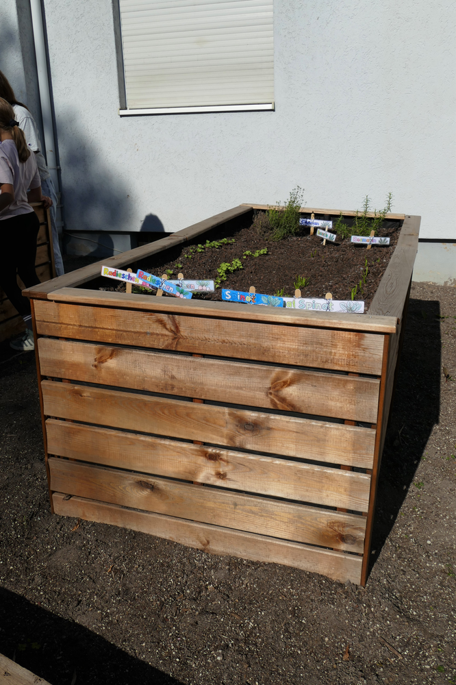 Ein Hochbeet mit verschiedenen kleinen Pflanzen und beschrifteten Schildern steht vor einer Hauswand, während einige Personen im Hintergrund zu sehen sind.
