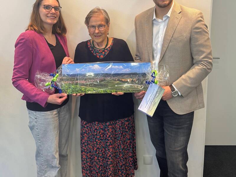 Drei Personen stehen nebeneinander in einem Raum, wobei die beiden äußeren Personen gemeinsam ein verpacktes, rechteckiges Geschenk halten.