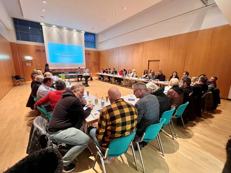 In einem Konferenzraum sitzen Menschen an einem großen U-förmigen Tisch und hören einer Präsentation zu.