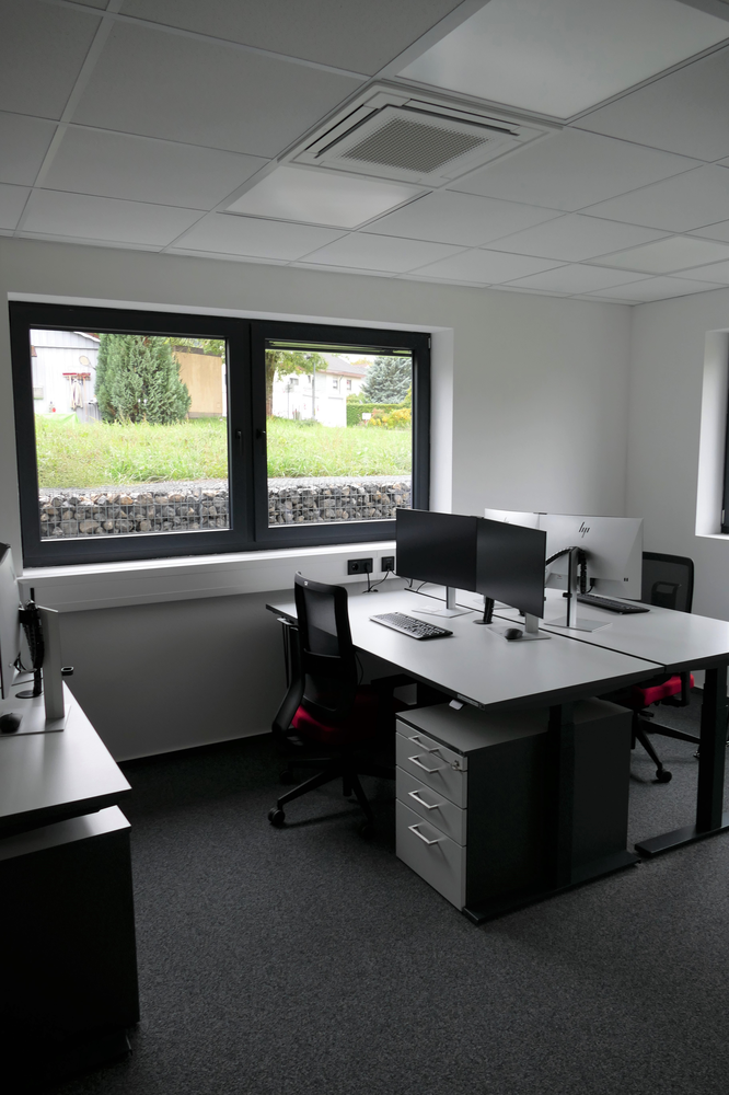 Ein modernes, helles Büro mit zwei Arbeitsplätzen, Computern und einem großen Fenster mit Blick nach draußen ist zu sehen.