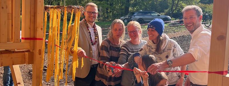 Eine Gruppe von Menschen, darunter Erwachsene und Kinder, steht fröhlich unter einem Holzgerüst und schneidet ein rotes Band durch.