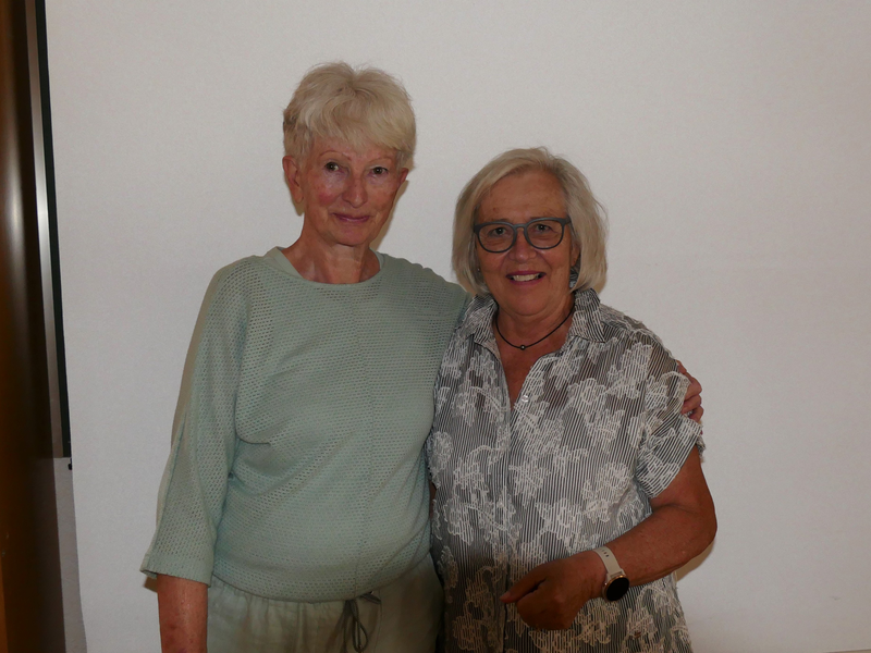 Zwei ältere Frauen stehen nebeneinander und lächeln in die Kamera, wobei eine Frau die andere freundlich mit dem Arm umarmt.