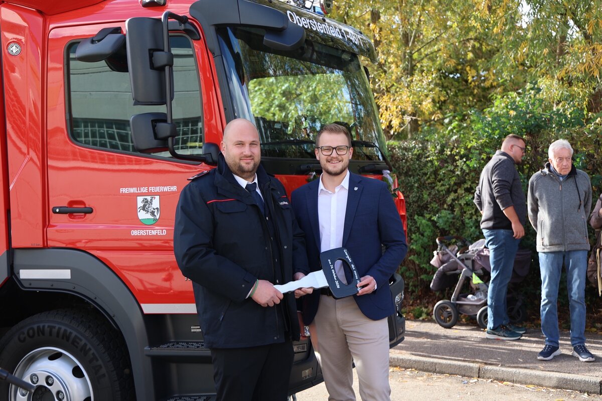 Zwei Männer stehen vor einem roten Feuerwehrauto, von dem einer ein Geschenk oder eine Auszeichnung in der Hand hält, während im Hintergrund weitere Personen auf einem Gehweg stehen.