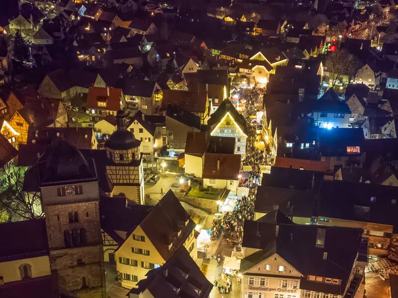 Eine nächtliche Luftaufnahme eines stimmungsvoll beleuchteten Dorfes, in dem ein belebter Weihnachtsmarkt zwischen zahlreichen Fachwerkhäusern stattfindet.